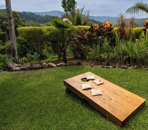 Un banc en bois avec des coussins, situé dans un jardin verdoyant, agrémenté de plantes tropicales et offrant une vue sur des collines.