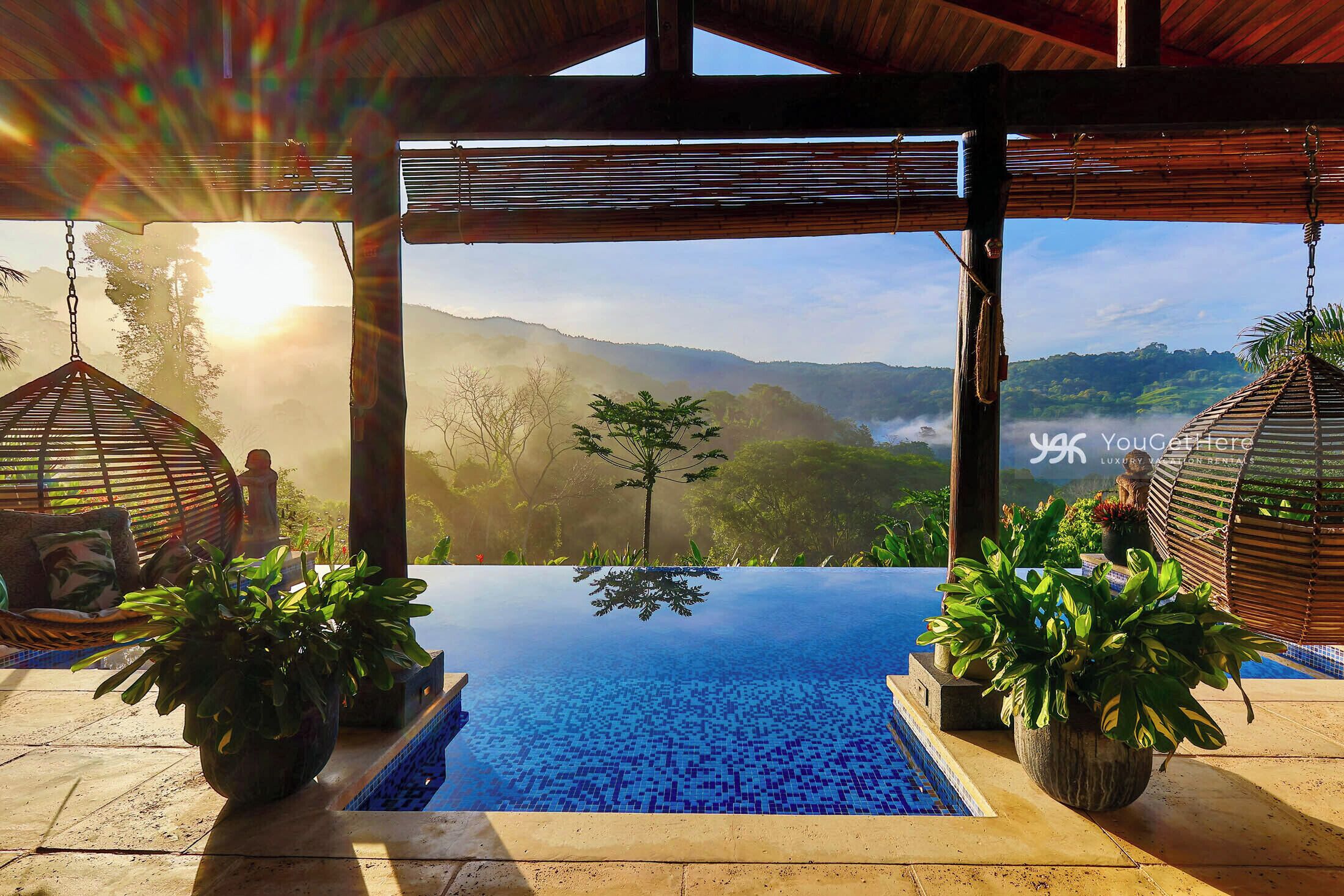 An infinity pool with a view of a mountainous landscape, surrounded by wooden structures and potted plants.