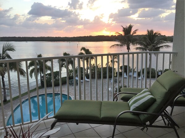 A balcony with a pool, lounge chairs, and a view of the sunset over the water.
