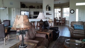 A spacious living room with a ceiling fan, leather sofas, a dining area with chairs, and a kitchen in the background.