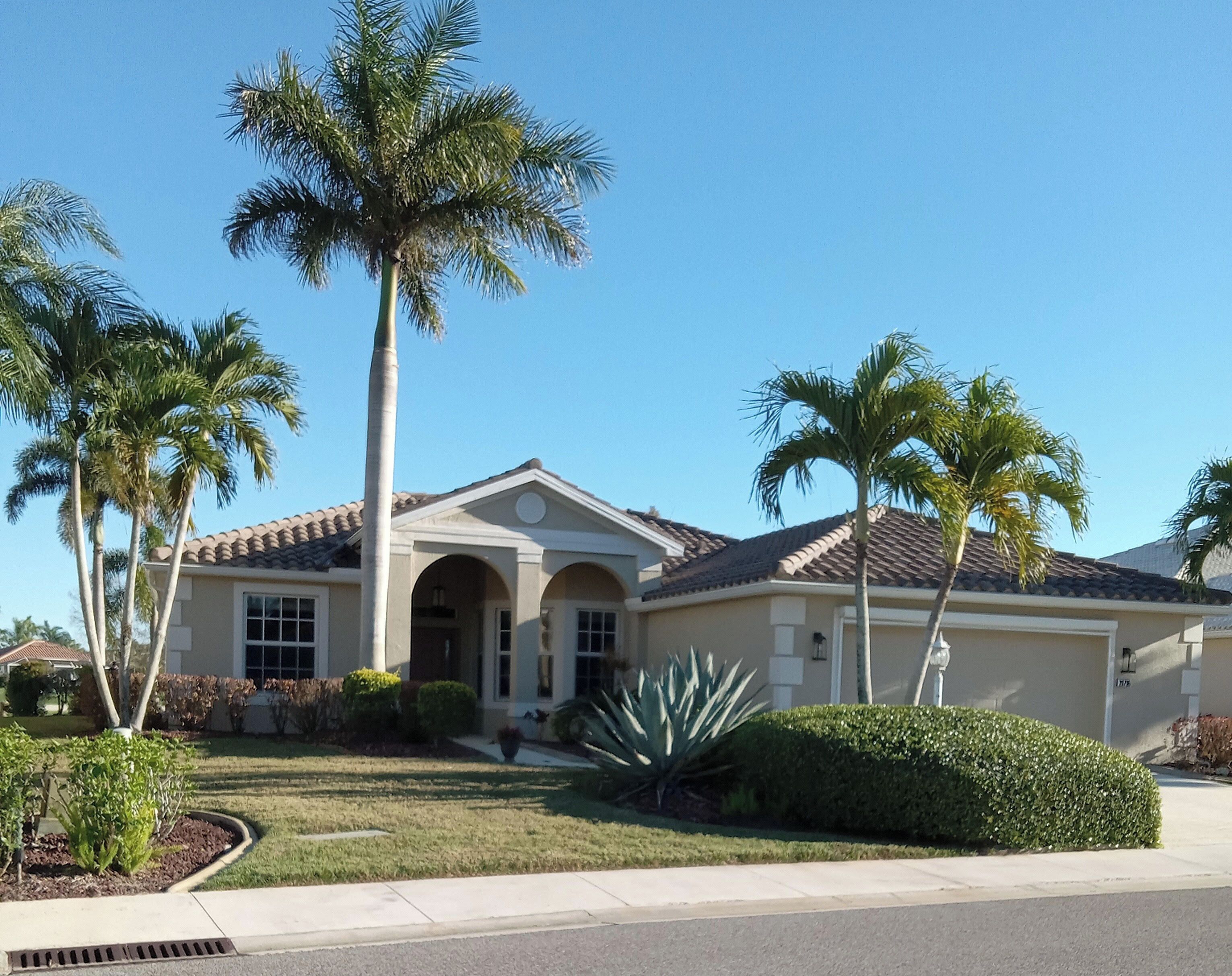 A two-story house with a tiled roof, a front entrance with an archway, and a well-maintained lawn with palm trees and shrubs.