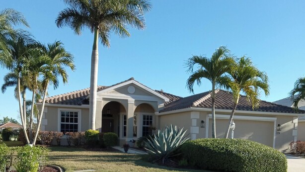 A two-story house with a tiled roof, a front entrance with an archway, and a well-maintained lawn with palm trees and shrubs.