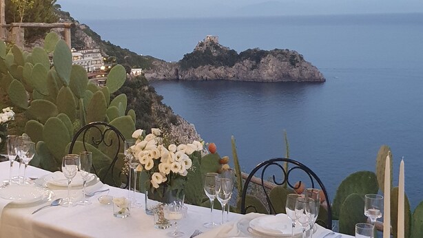 A table set for dining with a view of the sea and a rocky island in the background. - VILLA SHAH a place to listen to the sea in Amalfi Coast (Conca dei Marini)