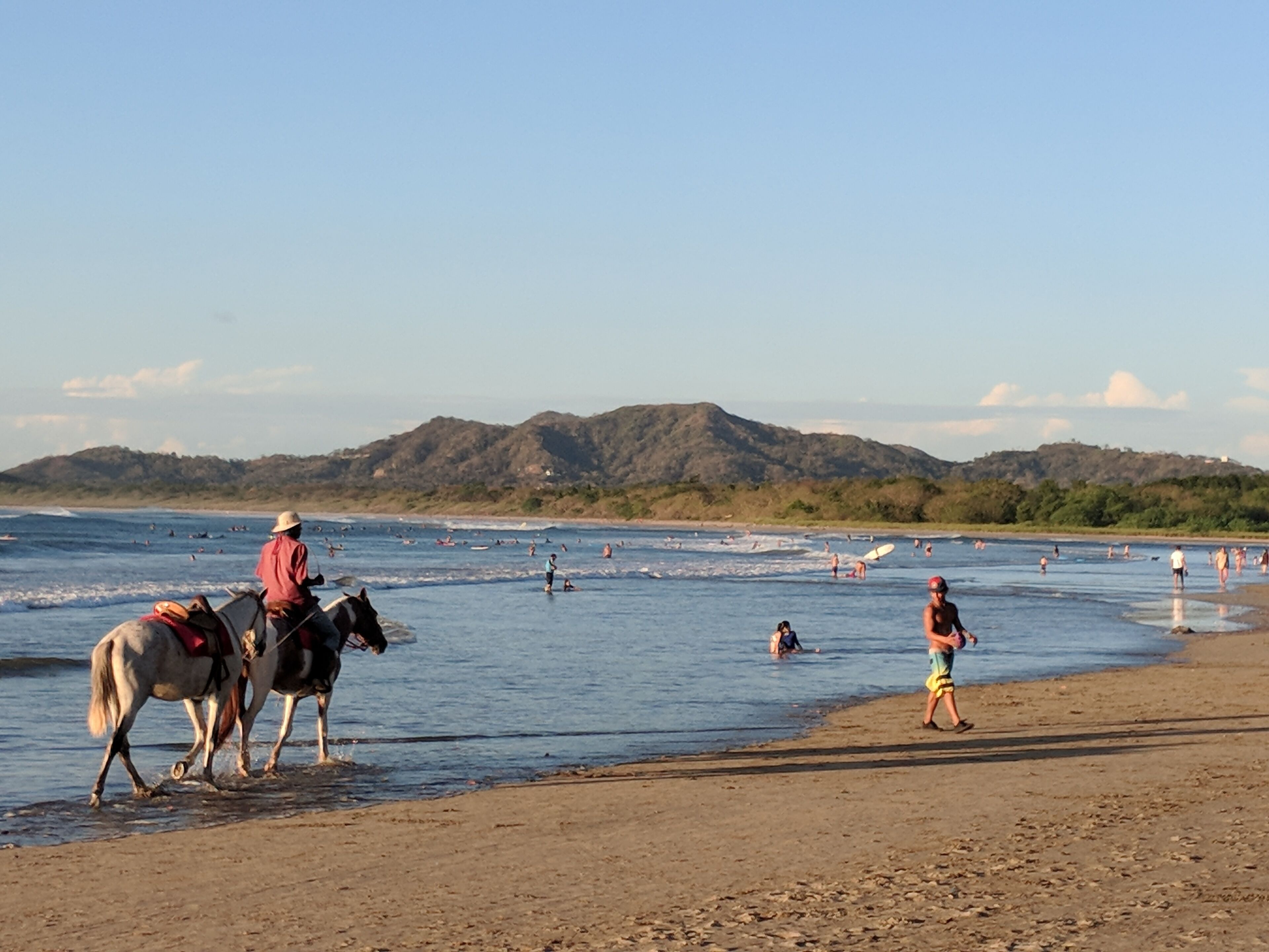 A beach with people and horses near the water, with hills in the background.
