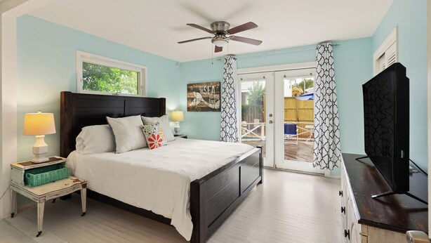 A bedroom with a bed, a ceiling fan, a TV, and a view to the outside.