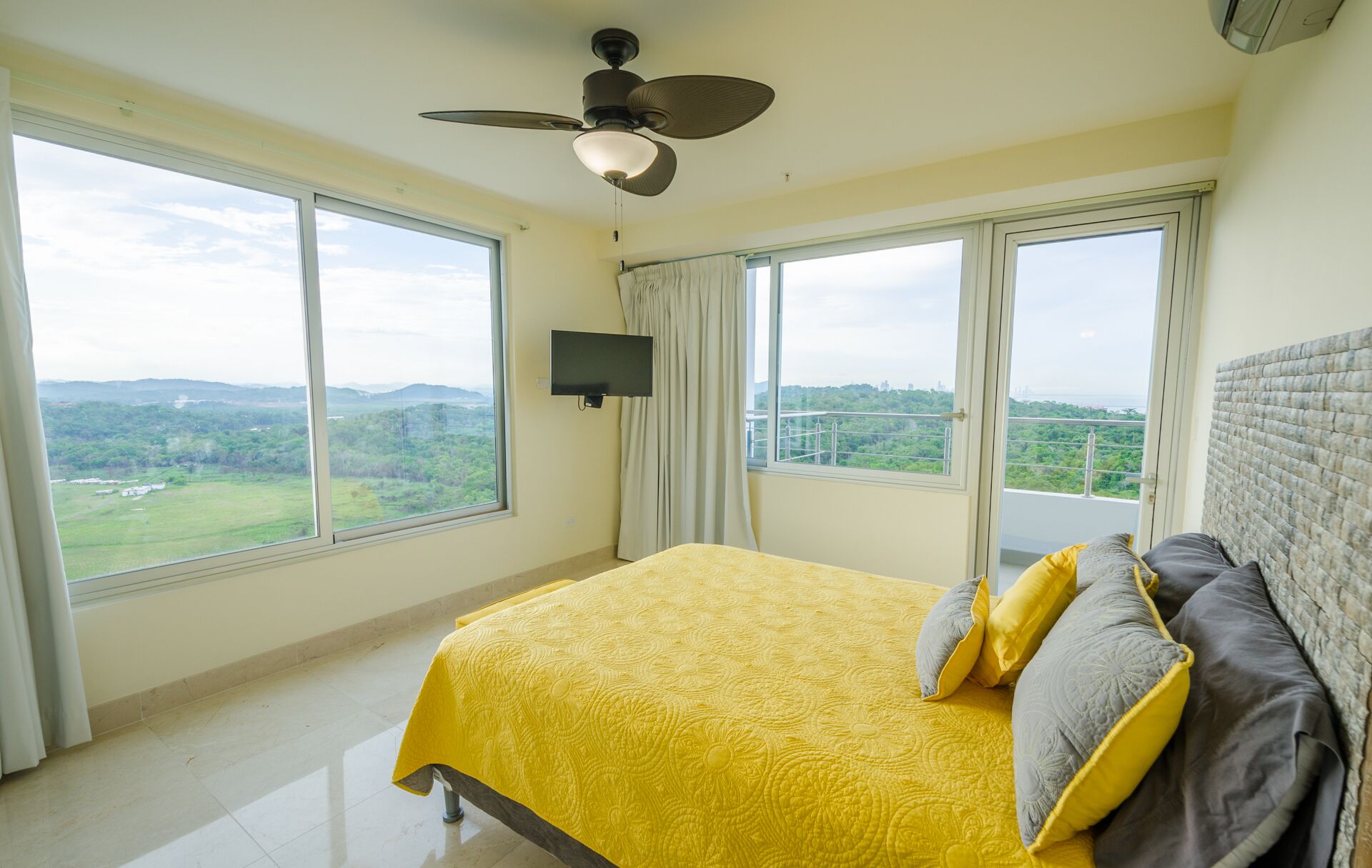 Un dormitorio con cama, ventilador de techo, televisor y ventanas amplias que ofrecen vistas al exterior.