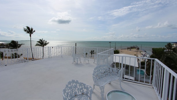 A balcony with white metal chairs and a glass table overlooking a beach with a pool and palm trees. - By The Sea ~ Islamorada Home with Ocean Views! (Islamorada)