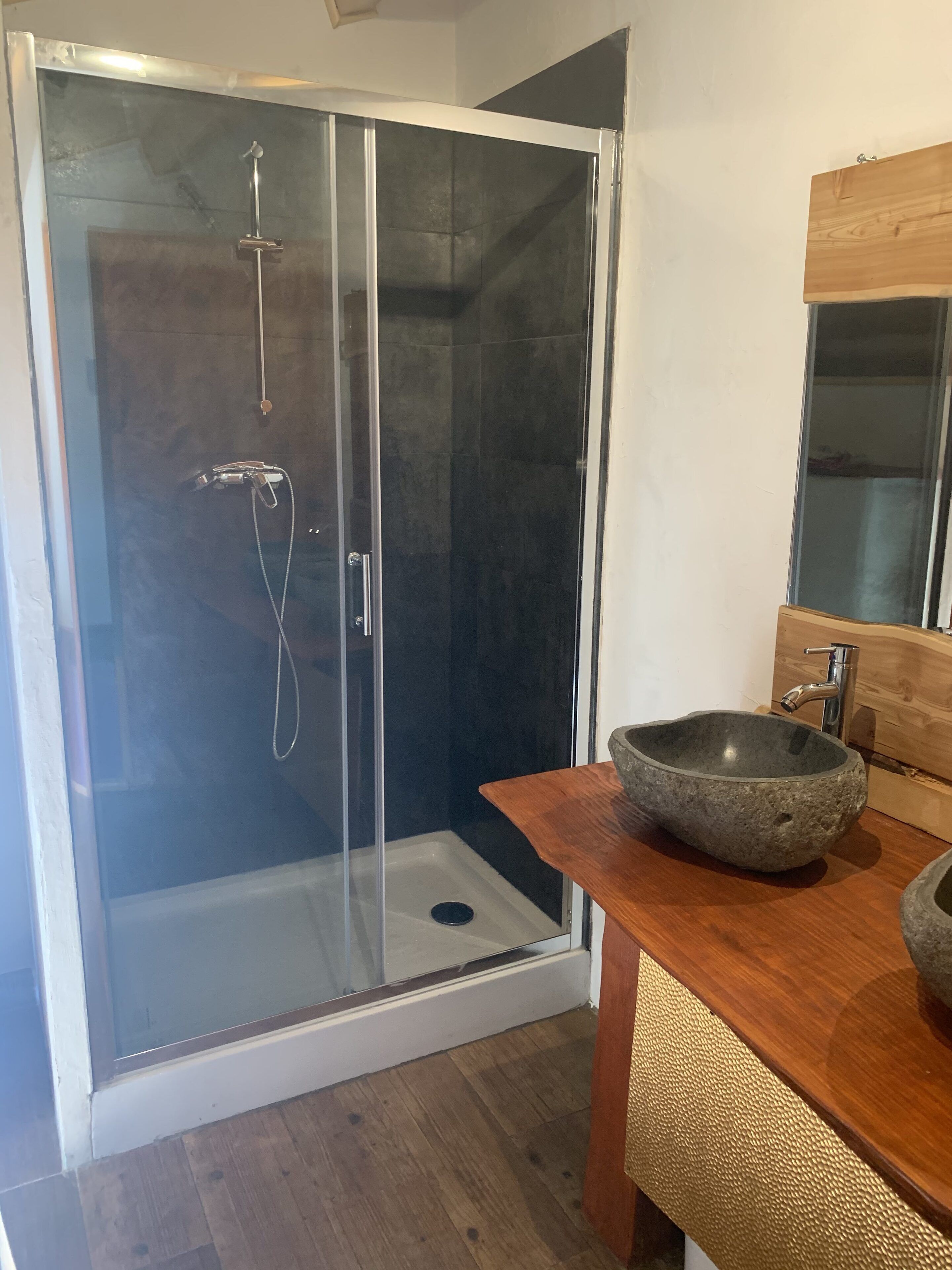 Un baño moderno con ducha envuelta en vidrio, tocador de madera con lavabo de piedra y un espejo grande sobre el lavabo.