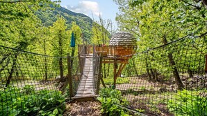 A wooden suspension bridge leading to a thatched-roof structure in a forested area with mountains in the background. - Les Ecrins de Belvedere (Belvedere)