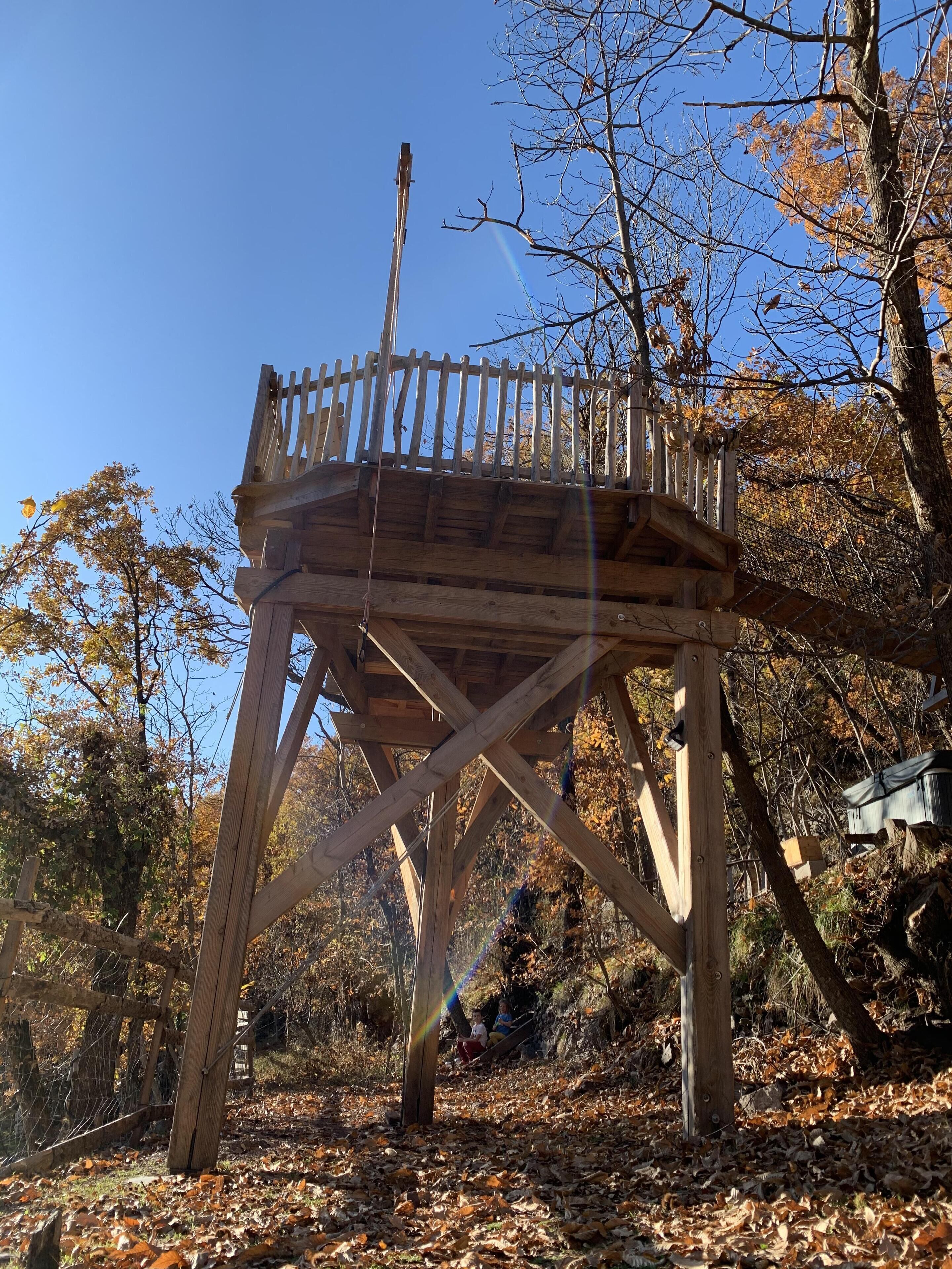A wooden observation tower with a metal railing, surrounded by trees and a clear sky.