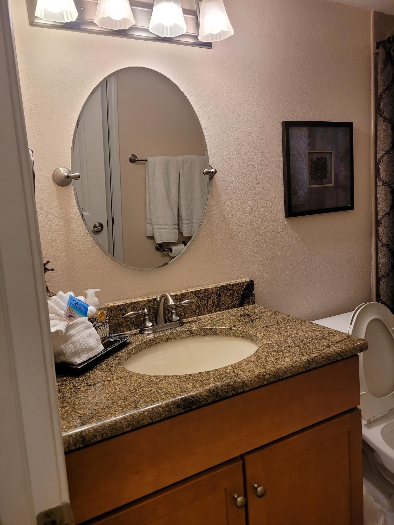 A bathroom with a granite countertop sink, a round mirror, a framed picture on the wall, and a towel hanging on the door.