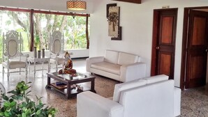 Amplia sala de estar con un sofá blanco, una mesita de centro y una estatua decorativa de Buda. Se aprecia una ventana grande con vistas a árboles y un techo de madera.