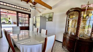 Un comedor con una mesa de vidrio, sillas de madera, ventilador de techo, refrigerador y un aparador antiguo.