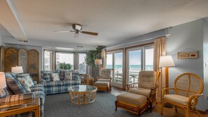 A spacious living room with a large window overlooking the beach, furnished with a sofa, armchairs, and a coffee table.