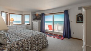 A bedroom with a bed, a dresser, a TV, and a view of the ocean through large windows.