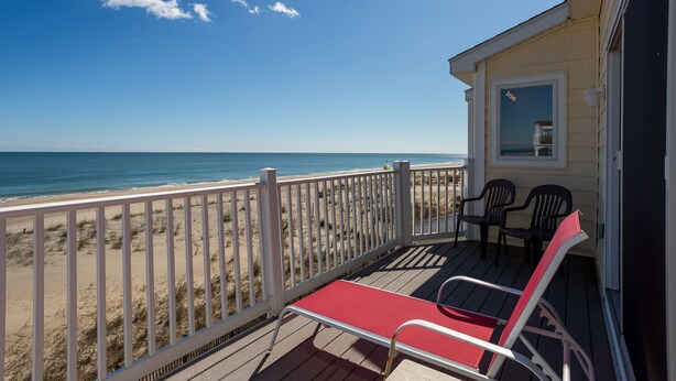 A beach house with a deck overlooking the ocean, featuring a red lounge chair and two black chairs.