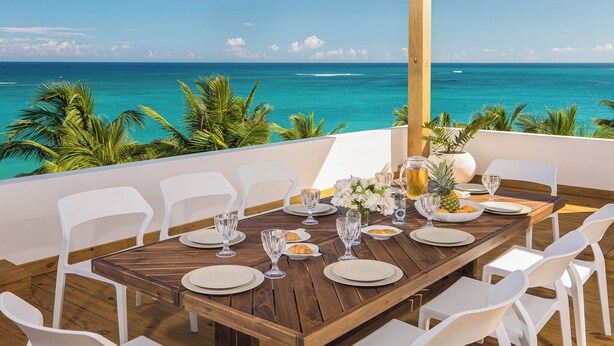 Une table en bois, dressée pour le repas, entourée de chaises blanches, avec vue sur un océan turquoise bordé de palmiers.