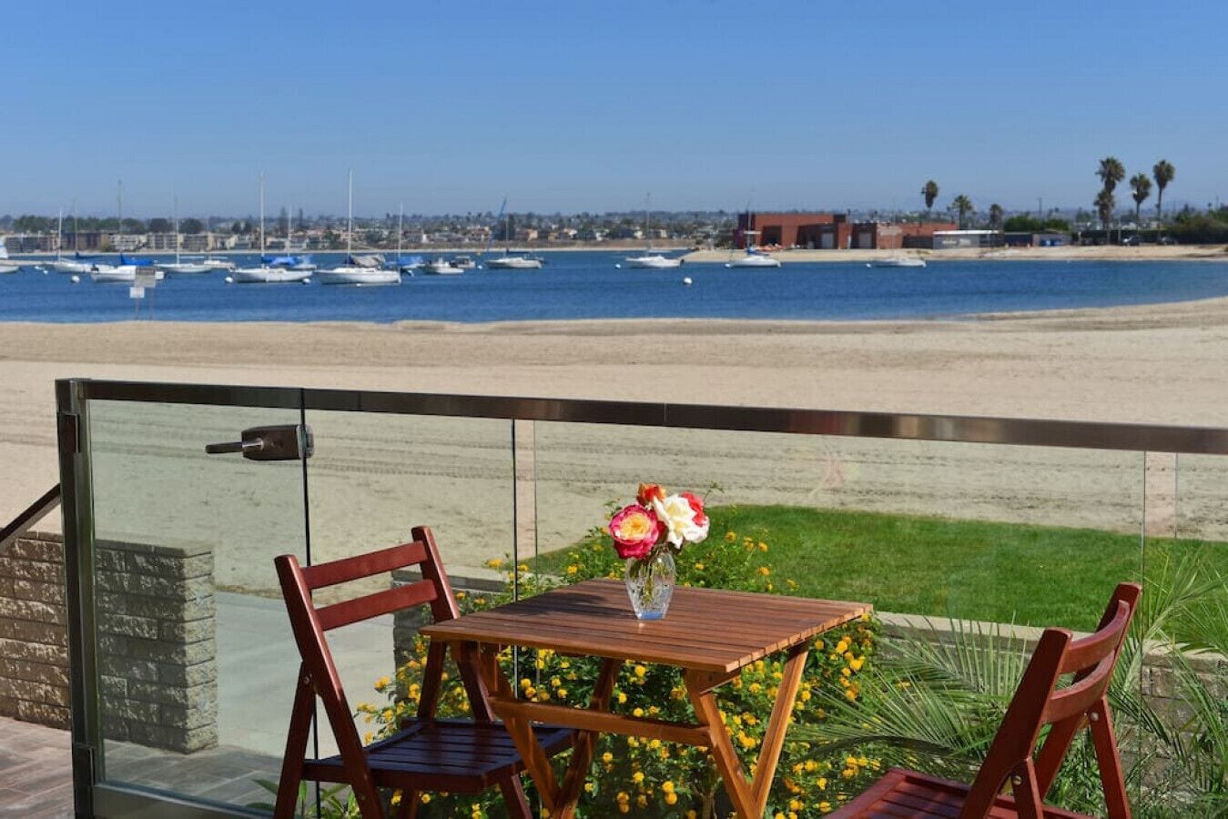 A wooden table with two chairs and a vase of flowers, overlooking a beach with boats and a distant shoreline.