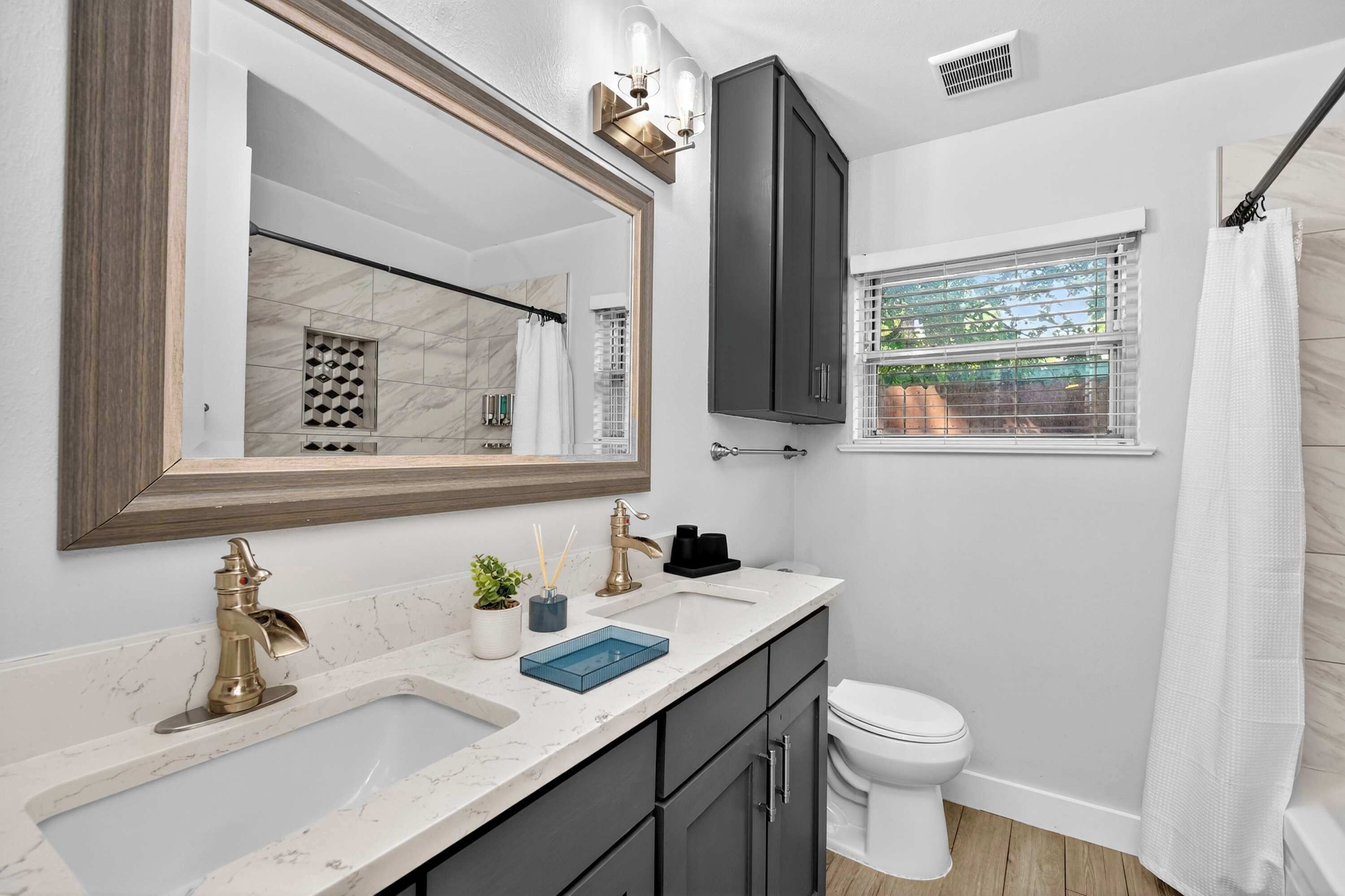 A bathroom with a large mirror, a sink with a gold faucet, a toilet, and a window with a shower curtain.