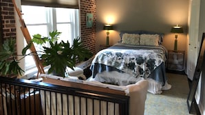 Ein Schlafzimmer mit Backsteinwand, Deckenventilator, einem Bett mit Blumenmuster auf der Bettwäsche, zwei Nachttischlampen und einer großen Pflanze.