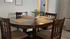 Un coin repas avec une table ronde en bois, entourée de quatre chaises dotées de coussins d’assise en tissu tressé. Une plante en pot est posée sur la table, et deux tableaux encadrés ornent le mur.