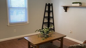 Une pièce avec une table en bois, une plante en pot, une échelle, une fenêtre avec des stores et une étagère sur laquelle est posé un panneau indiquant « MAISON ».