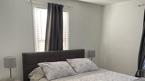 Une chambre à coucher comprenant un lit, des tables de chevet, une lampe, un ventilateur de plafond et une fenêtre avec des rideaux.