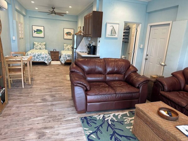 A cozy living room with a brown leather sofa, a wooden dining table, and two beds with patterned bedding.