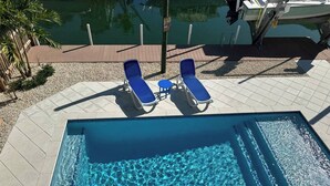 Una piscina rectangular con agua azul cristalina, rodeada por un solárium con baldosas. Hay dos tumbonas azules y una mesita.