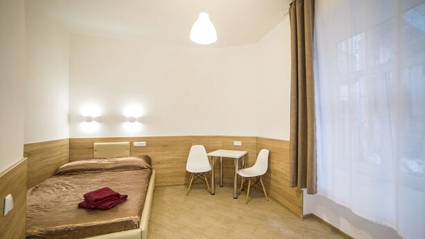A compact hotel room with a bed, a small table with two chairs, a wall-mounted light, and a window with curtains. - Smart Apartment Chornovola 21b (Lviv)