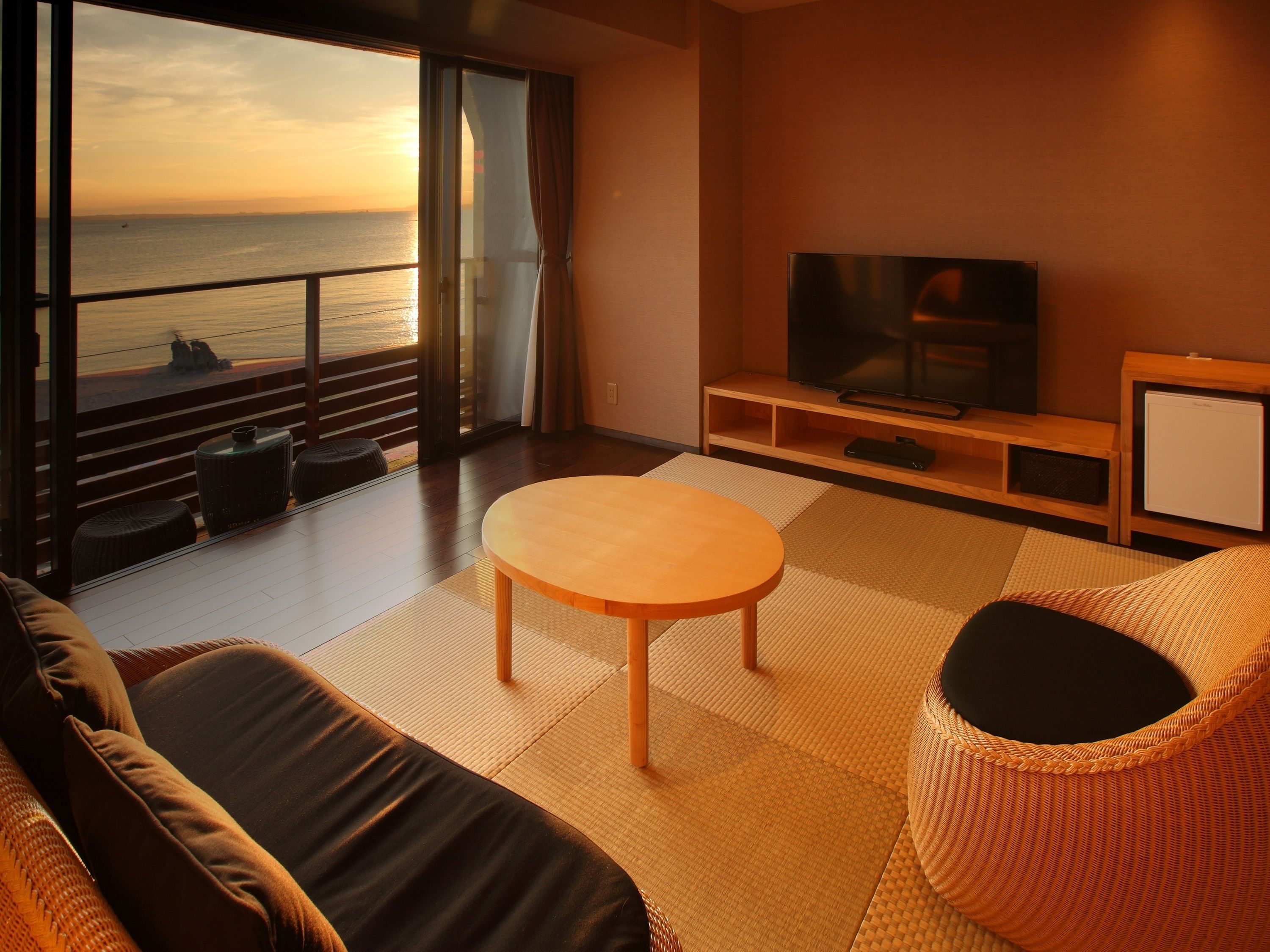 A modern living room with a large TV, comfortable seating, and a view of the ocean at sunset.