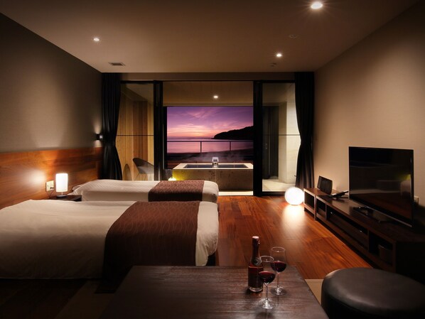 A hotel room with two beds, a TV, a table with wine glasses, and a view of the sunset. - Beachside Onsen Resort Umi (Kyonan)