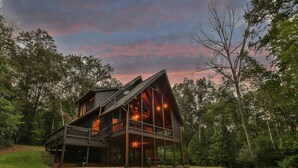 A large, multi-level cabin with a prominent front porch and multiple windows, surrounded by a forested landscape.