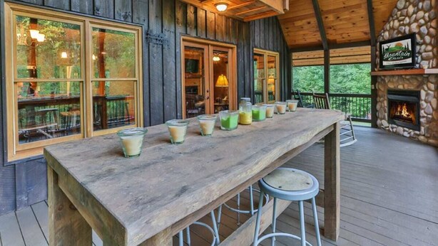 A wooden deck with a long table and stools, a stone fireplace, and large windows overlooking a forested area.