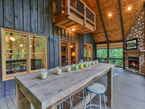 A wooden deck with a long table and stools, a stone fireplace, and large windows overlooking a forested area. - Southern Grace by Mountain Oasis | Creekfront Trout Fishing Cabin in McCaysville Near Blue Ridge (McCaysville)