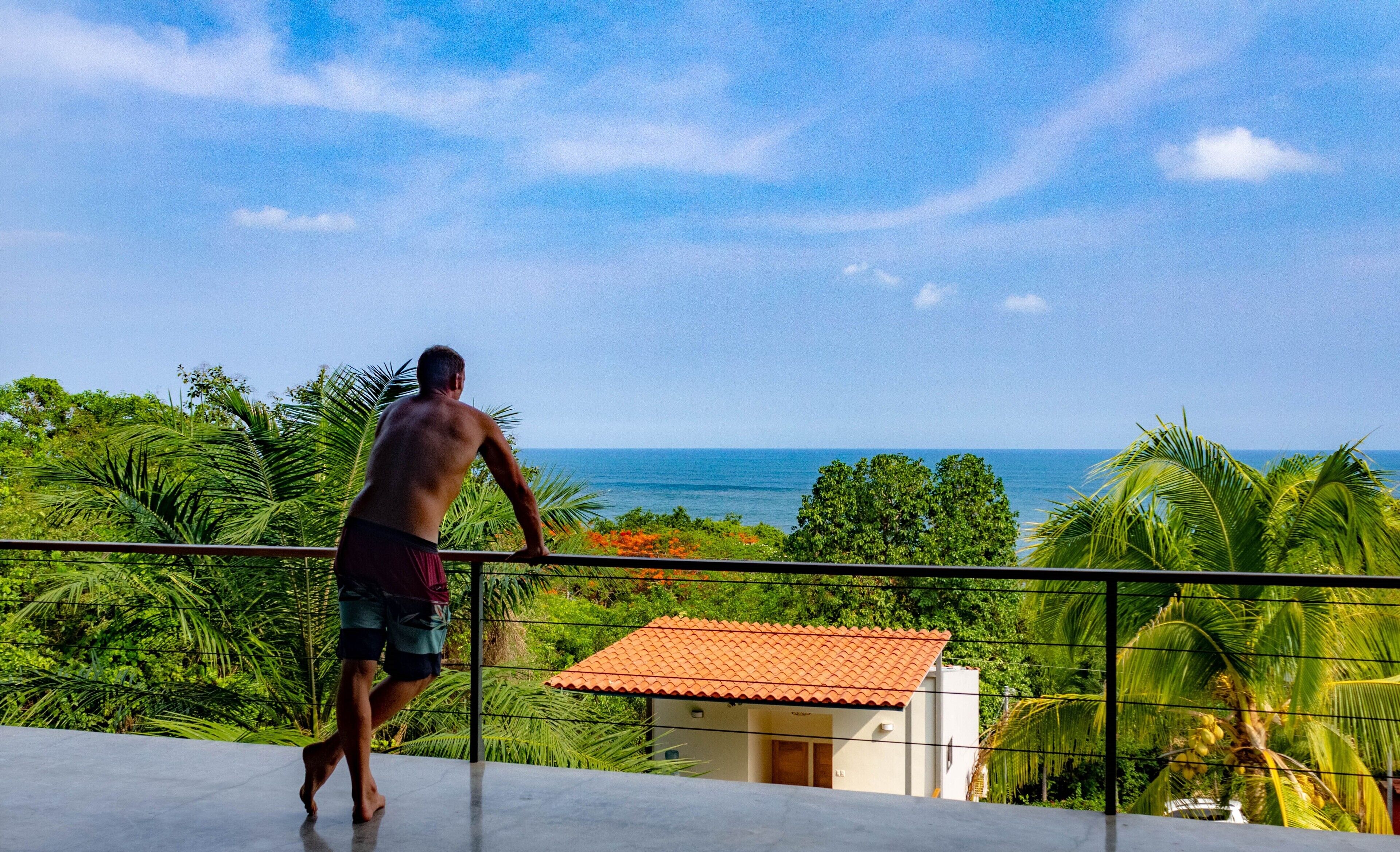 A man walking on a balcony overlooking a tropical landscape with a view of the sea.