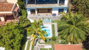 Une maison à deux étages avec une piscine, entourée de palmiers et d’un jardin.