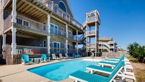 A pool area with lounge chairs and a building with multiple balconies. - Oceanfront Dog Friendly Home w/ Pool, Hot Tub, Game Room, Direct Beach Access (Waves)