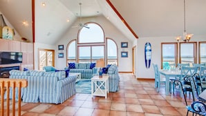 A spacious living area with a large window overlooking the sea, a ceiling fan, and a dining area with light blue chairs and tables. - Oceanfront Dog Friendly Home w/ Pool, Hot Tub, Game Room, Direct Beach Access (Waves)