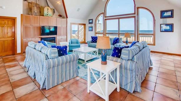 A spacious living room with striped sofas, a television, and large windows overlooking the sea. - Oceanfront Dog Friendly Home w/ Pool, Hot Tub, Game Room, Direct Beach Access (Waves)