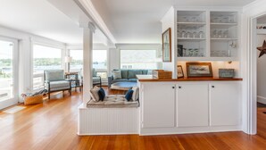 A spacious living area with a built-in seating area, a white cabinet with glassware, and a view of the outdoors. - Gardenwalk Crest, Elevated Harbor Views, Deck and Village Setting (Port Clyde)