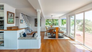 A spacious living area with a wooden floor, a dining table with chairs, a built-in bench with cushions, a staircase, and large windows offering a view of the outdoors. - Gardenwalk Crest, Elevated Harbor Views, Deck and Village Setting (Port Clyde)