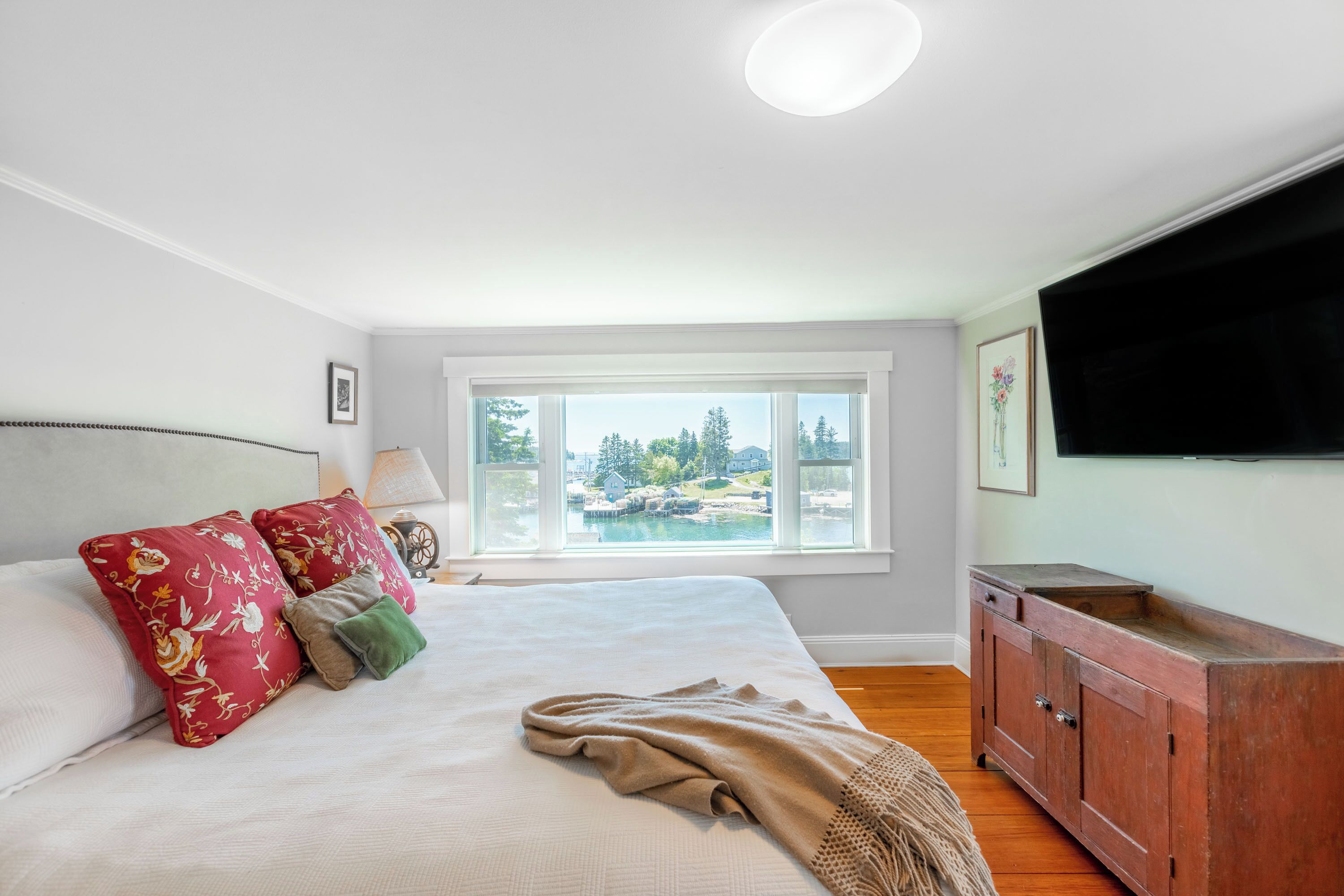 A bedroom with a large bed, a window with a view of a body of water, a wooden cabinet, and a flat-screen TV mounted on the wall.