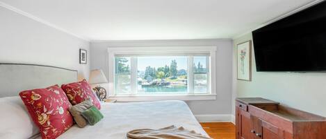 A bedroom with a large bed, a window with a view of a body of water, a wooden cabinet, and a flat-screen TV mounted on the wall.