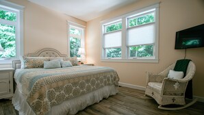 A bedroom with a large bed, a rocking chair, a television mounted on the wall, and large windows with blinds.