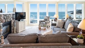 A living room with a stone fireplace, a television, a sofa, and large windows overlooking the ocean. - The Lighthouse, Whale Watching, Fireplace & Pool (Depoe Bay)