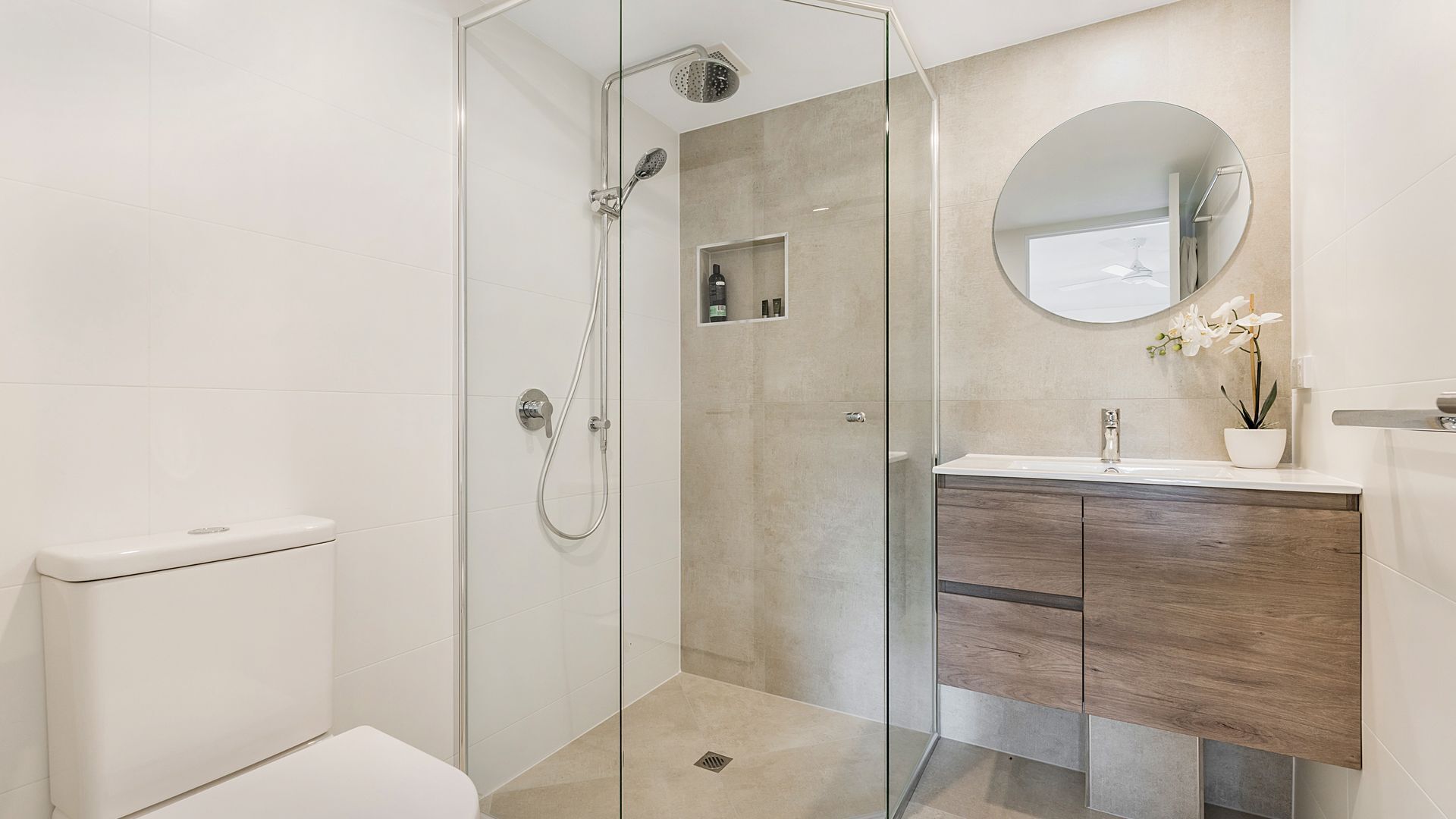 A modern bathroom with a glass-enclosed shower, a white toilet, a wooden vanity, and a round mirror.