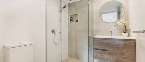 A modern bathroom with a glass-enclosed shower, a white toilet, a wooden vanity, and a round mirror.