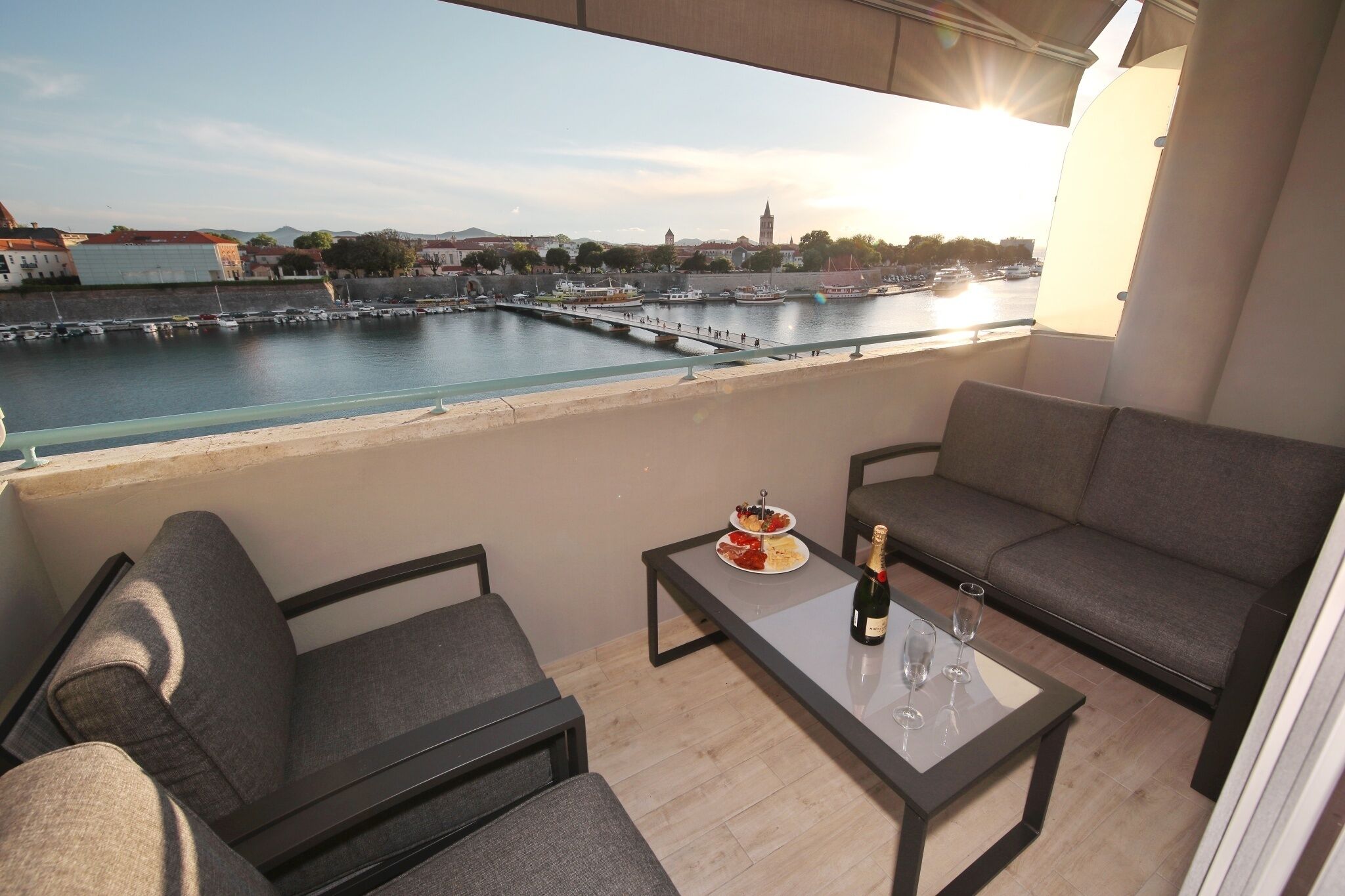 A balcony with a view of a harbor, a sofa, a table with a bottle and glasses, and a plate of food.