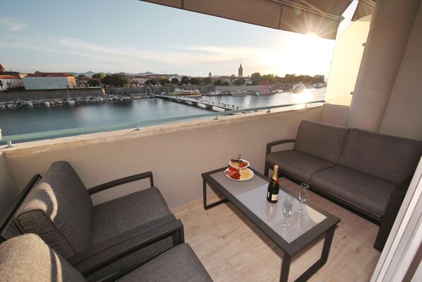 A balcony with a view of a harbor, a sofa, a table with a bottle and glasses, and a plate of food.
